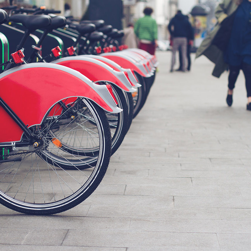 horizontal perspective view of city bike stand with row of red bicycles for rent on a city street and people passing by selective focus